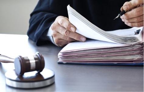 A person in a dark robe reviews legal documents at a desk, holding a pen. A wooden gavel rests nearby, suggesting a legal or judicial setting.